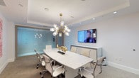 This conference room presents a refined and contemporary professional environment designed to facilitate productive meetings and collaborative work. The space centers around a large, glossy white conference table with rounded edges, comfortably accommodating multiple white upholstered ergonomic chairs with chrome bases, lending a clean and cohesive aesthetic throughout.

A standout design feature is the sculptural gold sputnik chandelier suspended from the ceiling, composed of clear glass globe bulbs on brass arms, serving as both a functional light source and an artistic focal point. Recessed ceiling lighting complements the chandelier, ensuring the room is evenly and warmly illuminated.

One wall features a wall-mounted flat-screen display, ideal for video conferencing and presentations, backed by a sleek low-profile credenza in a neutral finish. A full-height glass partition on the opposite side provides visual connectivity to adjacent spaces while maintaining acoustic separation.

Two large-scale abstract paintings in soft pink and blush tones adorn another wall, introducing warmth and a curated artistic sensibility to the otherwise neutral palette of white and greige. Neutral carpet flooring completes the professional yet inviting ambiance of this well-appointed meeting space.