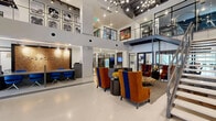 Located at 3700 Ross Avenue in Dallas, Texas, this expansive lobby for The Academic is a thoughtfully designed communal space that makes a striking first impression. The centerpiece of the space is a soaring double-height atrium that floods the interior with an airy, open ambiance, accentuated by large floor-to-ceiling windows that draw in natural light. Polished terrazzo flooring runs throughout, lending a clean, modern aesthetic that ties the entire space together.

A bold, large-scale mural in vibrant shades of blue, gold, and graphite commands attention along the upper wall, infusing the space with artistic energy. An industrial-style steel staircase with cable railings rises alongside a reclaimed wood accent wall, connecting the main level to the mezzanine above. The reception area features a textured cork feature wall backlit for warmth, paired with a dark wood desk and cobalt blue seating.

Adjacent to the reception, a neatly organized mailroom wall provides resident mailboxes beneath a stylishly lit ceiling with exposed black ductwork. A comfortable lounge area with richly upholstered orange and blue wingback chairs invites residents to gather and relax. Glass-partitioned rooms along the corridor offer additional functional spaces, while curated artwork displayed throughout adds cultural character to this well-appointed residential lobby.