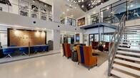 Located at 3700 Ross Avenue in Dallas, Texas, this expansive lobby for The Academic is a thoughtfully designed communal space that makes a striking first impression. The centerpiece of the space is a soaring double-height atrium that floods the interior with an airy, open ambiance, accentuated by large floor-to-ceiling windows that draw in natural light. Polished terrazzo flooring runs throughout, lending a clean, modern aesthetic that ties the entire space together.

A bold, large-scale mural in vibrant shades of blue, gold, and graphite commands attention along the upper wall, infusing the space with artistic energy. An industrial-style steel staircase with cable railings rises alongside a reclaimed wood accent wall, connecting the main level to the mezzanine above. The reception area features a textured cork feature wall backlit for warmth, paired with a dark wood desk and cobalt blue seating.

Adjacent to the reception, a neatly organized mailroom wall provides resident mailboxes beneath a stylishly lit ceiling with exposed black ductwork. A comfortable lounge area with richly upholstered orange and blue wingback chairs invites residents to gather and relax. Glass-partitioned rooms along the corridor offer additional functional spaces, while curated artwork displayed throughout adds cultural character to this well-appointed residential lobby.