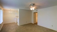 This single-story space presents a practical, open layout with a seamless connection between the main living area and the kitchen. The living room is finished with wall-to-wall carpeting in a neutral beige tone, providing a comfortable underfoot feel throughout the space. A ceiling fan with an integrated light fixture is centrally mounted on the textured popcorn ceiling, offering both air circulation and ambient lighting.

The walls are painted in a clean, off-white tone, creating a bright and neutral backdrop that complements the overall simplicity of the space. A partial wall or breakfast bar separates the living area from the kitchen, which is visible in the background and features white cabinetry and appliances. A hallway doorway is visible along one wall, suggesting additional rooms beyond the main living space.

The HVAC vent integrated into the ceiling indicates a central air system, contributing to year-round climate comfort. The straightforward design and neutral finishes make this space highly adaptable, providing an easy foundation for a variety of interior styles and furnishing arrangements.