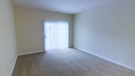 This single-floor space presents a straightforward and functional layout with a clean, neutral design. The main living area is finished with wall-to-wall carpet in a warm beige tone, providing a soft and comfortable underfoot feel throughout the room. The walls are painted in a light, neutral hue that contributes to a bright and open atmosphere.

A prominent sliding glass door serves as a key architectural feature, fitted with full-length vertical blinds that allow for adjustable natural light control. When open, the blinds reveal an exterior view, and the door provides convenient access to an outdoor area. The natural light filtering through the blinds brightens the interior, enhancing the sense of spaciousness.

The room includes standard white baseboards that offer a clean contrast against the carpeted floor, and a ceiling vent indicates a central HVAC system for climate control. The textured ceiling adds subtle depth to the space. Electrical outlets are positioned along the walls, offering practical placement for furnishings and electronics. Overall, the space maintains a simple, move-in-ready quality with a neutral palette suited to a variety of interior styles.