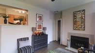 This single-story space features an inviting living area with warm, neutral-toned walls that create a welcoming atmosphere. The room is anchored by rich hardwood-style flooring, which flows seamlessly throughout and adds a sense of warmth and continuity. A ceiling fan overhead provides both functional airflow and a subtle design element.

A standout feature is the fireplace situated along one wall, framed in a clean, contemporary tile surround, serving as a natural focal point for the living area. Adjacent to the fireplace, a pass-through window with a ledge connects to the kitchen, offering a glimpse of cabinetry and décor beyond, while also allowing light to flow between the two spaces.

The living area is stylishly appointed with a dark, multi-drawer console cabinet topped with decorative lamps and amber-toned vases, complemented by two framed artwork pieces mounted above in warm earth tones. A striped accent chair adds texture and character to the seating arrangement. A door near the back wall leads to a closet or additional room, hinting at further functionality within the layout. Overall, the space balances comfort and style with thoughtful design details.