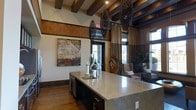 This thoughtfully designed single-floor space blends rustic charm with modern sophistication, creating a warm and inviting atmosphere. The interior is anchored by striking exposed dark wood ceiling beams that add architectural depth and character to the open layout. Dramatic woven pendant light fixtures hang from above, serving as bold focal points that complement the earthy, organic design palette.

The kitchen features a generously sized island topped with light-veined marble-style stone, offering ample surface space and a built-in sink. Dark cabinetry beneath the island provides a rich contrast to the lighter countertop. The floors throughout are finished in warm hardwood, reinforcing the cohesive natural aesthetic.

Transitioning into the living area, the space opens up to reveal comfortable seating arrangements bathed in natural light streaming through large windows dressed in flowing neutral drapes. Curated artwork and decorative accents, including a large mixed-media canvas and carved wood wall décor, add layers of visual interest.

A textured decorative band runs along the upper portion of the walls, adding an additional design element that ties the rustic and refined elements together. Overall, the space exudes a welcoming, upscale ambiance with a strong sense of warmth and intentional design throughout.