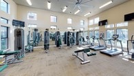 This single-floor fitness center is designed to support a comprehensive workout experience, combining thoughtful architecture with a full range of exercise equipment. The space features soaring ceilings with clerestory windows positioned along the upper walls, allowing abundant natural light to pour in from multiple angles and creating an open, energizing atmosphere. Large lower windows along one wall offer views of the surrounding outdoor environment, further enhancing the bright and welcoming ambiance.

The cardio zone is outfitted with multiple treadmills, elliptical trainers, upright bikes, and a recumbent bike, all positioned facing a bank of wall-mounted flat-screen televisions for entertainment during workouts. Overhead, a large ceiling fan and recessed lighting fixtures ensure consistent air circulation and illumination throughout.

The strength training area includes a variety of cable and resistance machines with green-accented benches, a dedicated free weight station with a full rack of dumbbells, and additional functional training equipment. Floor-to-ceiling mirrors line one wall, visually expanding the space and allowing users to monitor their form. Neutral-toned carpet tiles cover the floors throughout, providing comfort and durability. A posted fitness center rules board and a water station reflect the facility's attention to user convenience and safety.