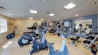 This fitness center offers a thoughtfully organized and fully equipped workout facility designed to accommodate a wide range of exercise routines. The space is bright and welcoming, illuminated by recessed and flush-mounted ceiling lights that ensure consistent, comfortable visibility throughout the room. The walls are finished in a clean combination of white and soft blue-gray tones, creating a calm and motivating atmosphere.

The cardio section is prominently featured, housing a row of treadmills, elliptical trainers, and stationary bikes — all from Life Fitness, a recognized brand in commercial fitness equipment. Each cardio machine is equipped with an individual monitor, allowing users to track workout metrics or enjoy entertainment during their session. The equipment is arranged in an orderly fashion, maximizing the use of floor space while maintaining ease of movement between machines.

Toward the back of the room, a dedicated strength training area provides additional workout options, including cable machines and weight stations. Motivational artwork adorns the walls, contributing to an energetic yet focused environment. The light-colored flooring ties the space together, keeping the overall aesthetic clean and professional. This fitness center is well-suited for users of all fitness levels.