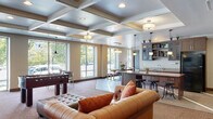 This single-floor community amenity space is thoughtfully designed to offer a variety of functional areas for gathering, recreation, work, and wellness. The centerpiece is a generously sized lounge area distinguished by elegant coffered ceilings, warm neutral tones, and an abundance of natural light streaming through large floor-to-ceiling windows that frame views of the surrounding greenery. Comfortable tufted leather sofas and upholstered seating are arranged to encourage both relaxation and socializing, while a foosball table adds a recreational touch. Adjacent to the lounge, a full kitchen features dark wood cabinetry, granite countertops, a full-size refrigerator, a sink, and bar-height seating along an island — ideal for events and casual gatherings. The business office is furnished with rich mahogany desks and cabinetry, multiple computer workstations, and coffered ceiling detailing consistent with the rest of the space, providing a professional yet welcoming environment. The fitness room is equipped with cardio machines and strength training equipment set against a bold accent wall, accompanied by a ceiling fan for comfort. The entryway features tile flooring with inlay detailing, wood-trimmed doors, and a tray ceiling, setting a polished tone upon arrival.
