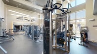 Located at Spring Plaza Drive in Spring, Texas, this modern fitness center offers a generously sized, single-floor workout space designed to accommodate a full range of exercise needs. The facility's most striking architectural feature is its soaring ceiling height, which lends an open, airy feel throughout the entire space. Tall, floor-to-ceiling windows line the perimeter, flooding the interior with natural light and offering views of the outdoor surroundings, including a glimpse of a pool area and landscaping beyond.

The design incorporates stacked stone accent columns that add a refined, contemporary aesthetic, complemented by neutral tones and polished surfaces. Large wall-mounted mirrors extend the visual depth of the space, while mounted flat-screen televisions provide entertainment during workouts.

The equipment layout is thoughtfully organized, with a dedicated cardio zone featuring premium treadmills — including Woodway models — StairMasters, ellipticals, stationary bikes, and cycling machines positioned along the windowed wall. The strength training area is equally well-appointed, offering cable machines, Smith machines, free weights including a full dumbbell rack, weight benches, and a variety of selectorized strength machines. Rubber flooring throughout ensures safety and durability. Ceiling fans contribute to a comfortable workout environment year-round.