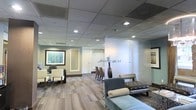 This well-appointed single-floor professional space offers a thoughtfully designed environment that balances functionality with refined aesthetics. The reception and waiting areas are furnished with contemporary seating, including plush upholstered chairs and a tufted bench in soft blue-gray tones, creating a welcoming and comfortable atmosphere for visitors. Glass partitions divide the space with a sense of openness while maintaining privacy, allowing light to flow freely between areas. Recessed lighting set within a drop ceiling provides consistent, warm illumination throughout. The flooring features a subtle, patterned carpet in neutral tones that complements the muted color palette of sage green and warm beige walls. A framed abstract artwork adds a touch of sophistication to the reception area, while a tall decorative vase with branch accents serves as an elegant focal point near the glass partition. Beyond the reception zone, a glimpse into an adjoining room reveals additional seating, suggesting a conference or meeting area. The overall ambiance is calm, professional, and polished, reflecting a space designed to make a strong first impression while remaining functional and inviting for daily business operations.