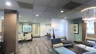 This well-appointed single-floor professional space offers a thoughtfully designed environment that balances functionality with refined aesthetics. The reception and waiting areas are furnished with contemporary seating, including plush upholstered chairs and a tufted bench in soft blue-gray tones, creating a welcoming and comfortable atmosphere for visitors. Glass partitions divide the space with a sense of openness while maintaining privacy, allowing light to flow freely between areas. Recessed lighting set within a drop ceiling provides consistent, warm illumination throughout. The flooring features a subtle, patterned carpet in neutral tones that complements the muted color palette of sage green and warm beige walls. A framed abstract artwork adds a touch of sophistication to the reception area, while a tall decorative vase with branch accents serves as an elegant focal point near the glass partition. Beyond the reception zone, a glimpse into an adjoining room reveals additional seating, suggesting a conference or meeting area. The overall ambiance is calm, professional, and polished, reflecting a space designed to make a strong first impression while remaining functional and inviting for daily business operations.
