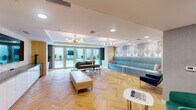 This contemporary amenity lounge is designed to impress, combining thoughtful layout with refined interior finishes throughout its single floor. The space is anchored by striking herringbone-patterned hardwood floors that run continuously, lending warmth and visual continuity to the entire area. Recessed lighting is complemented by multiple sculptural globe chandeliers that add a modern, artistic touch to the ceiling.

The lounge is divided into distinct yet open seating zones. A built-in banquette upholstered in teal velvet lines one wall, backed by a decorative Art Deco-inspired laser-cut panel that serves as a dramatic focal point. Complementary freestanding sofas in teal and warm brown tones, paired with round accent tables, create comfortable conversation areas throughout the room.

A large flat-screen television is mounted on a textured three-dimensional white tile wall, accompanied by an extended white cabinetry run with a stone-topped wet bar and integrated sink — ideal for entertaining. A beverage refrigerator is also built in below the counter.

Floor-to-ceiling glass French doors along one wall flood the space with natural light and open directly onto a landscaped outdoor terrace, seamlessly connecting interior and exterior. Additional accent chairs, a tufted bench, and a small chess table complete the social atmosphere of this well-appointed gathering space.