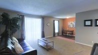 This single-floor space presents an open and functional layout that seamlessly connects the living and dining areas. The living room is generously sized, featuring plush wall-to-wall carpeting and a large sliding glass door dressed with vertical blinds and draped curtains, allowing for abundant natural light and easy outdoor access. A secondary window with coordinating curtains further brightens the space. The textured ceiling adds a classic architectural detail throughout.

Adjacent to the living area, the dining nook makes a visual statement with a warm orange accent wall that creates a cozy and inviting atmosphere. The space accommodates a dark wood dining table and chairs, complemented by a patterned area rug and colorful wall art, adding personality and depth to the room. Decorative elements such as branch accents and a vase provide stylish finishing touches.

The neutral taupe wall tones throughout the living area create a versatile backdrop, making it easy to personalize the space. Together, the open sightlines between the living and dining zones foster a connected, social atmosphere, while the distinct color palette of each area gives each zone its own defined character. This well-lit, single-level layout is designed for comfortable everyday living.