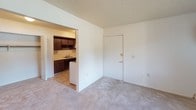 This single-floor unit offers a straightforward, functional layout across several distinct areas. The living area features neutral-toned carpet underfoot and white walls beneath a textured popcorn ceiling, creating a clean, blank-canvas atmosphere. A large window fitted with vertical blinds allows natural light to fill the space, while a baseboard heater runs along the wall. Adjacent to the living area, the kitchen is accessible through an open passageway and is equipped with dark walnut-toned cabinetry — both upper and lower — paired with light laminate countertops and a single-basin stainless steel sink. Tile flooring transitions the kitchen from the carpeted living space. The bedroom is a well-proportioned room with carpet, a window overlooking the outdoors, a baseboard heater, and a closet with louvered doors. The bathroom includes a full-size soaking tub and shower combination, a toilet, and a vanity with sink, all set on tile flooring with a textured ceiling above. Throughout the unit, the design is practical and neutral, offering a versatile foundation for personalization.