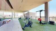 This expansive rooftop fitness facility combines open-air and covered workout environments, offering a dynamic training experience elevated above a bustling urban skyline. The space is organized across a generous rooftop level, where artificial turf flooring defines the primary workout zones, creating a functional yet visually striking surface throughout.

The covered sections feature clean-lined concrete columns and flush linear lighting embedded in smooth overhead panels, providing shade while maintaining an open, airy feel. Floor-to-ceiling glass walls along one interior-facing side allow natural light to penetrate the space and offer views into adjacent interior areas.

Equipment is thoughtfully distributed throughout, including air bikes, manual treadmills, cable machines, and sled trainers on the turf sections. A dedicated boxing and combat training area features hanging speed bags, an Octagon dumbbell rack, and striking equipment. Resistance tools such as sandbags and medicine balls are neatly organized on storage racks nearby.

The perimeter is lined with glass railings, offering unobstructed panoramic views of the surrounding city skyline and a resort-style pool deck below. The combination of sunlit open-air zones and sheltered covered areas makes this facility suitable for year-round training.