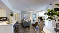 This well-appointed single-floor residence offers an open-concept design that integrates the living, dining, and sleeping areas into a cohesive and functional layout. Dark hardwood-style flooring runs throughout, providing a warm and grounding contrast to the crisp white ceilings and light gray accent walls.

The central living area is anchored by a comfortable gray sofa, complemented by an accent armchair and a stylish floor lamp, creating an inviting space for relaxation. A ceiling fan overhead adds both practicality and a finishing architectural touch. Wall art and carefully chosen décor lend the space a curated, modern sensibility.

Adjacent to the living area, a dining nook with bar-style seating connects seamlessly to what appears to be a kitchen space, making entertaining straightforward and convenient. A media console with a flat-screen television is positioned along one wall, completing the entertainment-focused living zone.

Natural light filters in through a wide window dressed with sheer white curtains, brightening the space and creating an airy atmosphere. A glimpse into the bedroom reveals a neatly appointed sleeping area, while a bathroom with a shower curtain is also visible, indicating a well-organized and efficiently designed floor plan. Overall, the space balances contemporary style with everyday comfort.