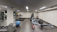 This fitness center offers a comprehensive workout environment designed to accommodate a wide range of exercise needs. The space is laid out across a single open floor, providing ample room to move between equipment zones without feeling congested. Durable rubber flooring covers the entire area, offering both safety and sound absorption throughout.

The cardio section includes multiple treadmills, elliptical trainers, a rowing machine, and a stationary bike, providing diverse options for aerobic training. A dedicated strength training area features several multi-station cable machines, ideal for a full range of resistance exercises. Free weight resources include a complete dumbbell rack and multiple adjustable benches for various pressing and accessory movements.

Functional training is also well represented, with stability balls and other accessories available for core and flexibility work. Large mirrors line portions of the walls, enhancing the sense of space while allowing users to monitor their form. Recessed fluorescent lighting ensures the entire area is brightly and evenly illuminated. A wall-mounted television provides entertainment, and a water cooler is conveniently accessible. A window along one wall introduces a degree of natural light into the space.