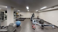 This fitness center offers a comprehensive workout environment designed to accommodate a wide range of exercise needs. The space is laid out across a single open floor, providing ample room to move between equipment zones without feeling congested. Durable rubber flooring covers the entire area, offering both safety and sound absorption throughout.

The cardio section includes multiple treadmills, elliptical trainers, a rowing machine, and a stationary bike, providing diverse options for aerobic training. A dedicated strength training area features several multi-station cable machines, ideal for a full range of resistance exercises. Free weight resources include a complete dumbbell rack and multiple adjustable benches for various pressing and accessory movements.

Functional training is also well represented, with stability balls and other accessories available for core and flexibility work. Large mirrors line portions of the walls, enhancing the sense of space while allowing users to monitor their form. Recessed fluorescent lighting ensures the entire area is brightly and evenly illuminated. A wall-mounted television provides entertainment, and a water cooler is conveniently accessible. A window along one wall introduces a degree of natural light into the space.