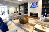 Located at 712 Promenade Place in Virginia Beach, VA, this single-floor community clubhouse offers a thoughtfully designed, multi-functional environment with a contemporary aesthetic throughout. The open-plan main gathering area greets visitors with warm wood-look flooring, white structural columns with transom detailing, and a striking globe chandelier that anchors the entryway. This central hub flows seamlessly into a comfortable lounge featuring a tray ceiling, built-in shelving, a fireplace with a stone surround, and a wall-mounted television — ideal for relaxed socializing. A well-appointed kitchen area includes granite countertops, a large island with bar seating, pendant lighting, open shelving, and stainless steel appliances, providing a functional and inviting space for residents. A dedicated conference room accommodates large group gatherings around a sleek table beneath a modern chandelier, with natural light filtering in through Roman shade-dressed windows. A smaller breakout room with a round glass table offers a more intimate setting for smaller meetings. The business center provides computer workstations and printing capabilities in a focused, quietly styled environment. A media room with dark walls, plush leather seating, and a large wall-mounted screen rounds out the amenities. A billiards area and fitness space further enhance the breadth of offerings in this well-rounded community facility.