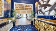 Located in Ocala, Florida, this beautifully appointed single-story clubhouse at 3491 SW 42nd St delivers an impressive blend of coastal-inspired design and upscale finishes throughout. The grand entrance immediately captures attention with striking cobalt blue hexagonal tile flooring, bold navy accent walls, and dramatic gold sculptural décor elements that set an unmistakably sophisticated tone.

The soaring coffered ceilings with warm wood inlays and recessed lighting define the open central gathering areas, which are bathed in natural light streaming through expansive floor-to-ceiling glass doors that lead directly to the pool area. A statement lounge area features a floor-to-ceiling mirrored tile fireplace flanked by stacked stone accents, a mounted TV, and curated seating with rich wood and gold-toned furnishings centered around a gleaming gold cocktail table.

The well-equipped kitchen showcases slate-blue cabinetry with brass hardware, quartz countertops, a decorative mosaic tile backsplash, stainless steel appliances including full-size refrigerators, and marble-look tile flooring. Additional interior corridors connect secondary gathering rooms and multi-purpose spaces, all finished with consistent marble-look tile, shiplap-style wainscoting, and tasteful decorative accents. A dedicated meeting or event area with a conference table and digital display rounds out the space, offering both function and style for community use.