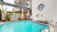 Located in the desirable Brentwood neighborhood of Los Angeles, this outdoor pool area offers a well-appointed communal retreat defined by its classic architectural character. The centerpiece is a generously sized freeform swimming pool with vivid turquoise water, framed by a smooth pool deck furnished with lounge chairs and casual seating—ideal for relaxing in the Southern California sun.

A covered pergola structure with white columns and a blue-trimmed roofline extends over a portion of the deck, providing shaded outdoor space and lending a resort-like atmosphere to the area. The surrounding multi-story building features white-painted brick walls punctuated by distinctive circular porthole-style architectural accents stacked vertically, adding a nautical design flair that sets this property apart.

Lattice privacy fencing, potted plants, and mature greenery create a lush, semi-enclosed environment that feels both private and inviting. Blue metal railings along the exterior stairways complement the building's consistent color palette. The pool area is enclosed by iron fencing and bordered by tall hedges, ensuring a sense of seclusion while remaining open to the sky. The overall ambiance is bright, clean, and welcoming, reflecting the relaxed yet polished character of its Los Angeles setting.