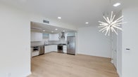 This single-floor space showcases a clean, modern aesthetic defined by an open-concept design that seamlessly connects the kitchen and living areas. The kitchen is outfitted with flat-front white cabinetry, dark stone countertops, and a full suite of stainless steel appliances, including a gas range, French door refrigerator, built-in oven, and dishwasher. A stainless steel wall-mounted range hood serves as a functional centerpiece above the cooktop. The L-shaped kitchen layout maximizes efficiency while maintaining an unobstructed flow into the adjacent living space. Light-toned wide-plank hardwood flooring extends throughout, contributing to a bright and airy atmosphere. Recessed lighting is installed throughout the ceiling, providing consistent, even illumination. A striking starburst chandelier adds a bold decorative focal point to the open living area, contrasting elegantly with the otherwise minimalist design. Crisp white walls and a neutral color palette enhance the sense of openness, making the space feel expansive and well-lit. The overall ambiance is contemporary and refined, ideal for modern living.