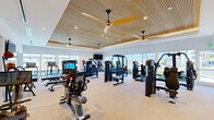 Located at 401 SE Central Pkwy in Stuart, Florida, this single-level fitness center offers a generously sized, thoughtfully designed space dedicated to health and wellness. The facility's interior is defined by a standout tray ceiling clad in warm slatted wood paneling, accented by recessed lighting and large paddle ceiling fans that contribute to both aesthetic appeal and air circulation throughout the space.

The main workout area is expansive and open, accommodating a comprehensive selection of commercial-grade equipment. Cardio machines—including treadmills, ellipticals, stationary bikes, and rowing machines—are positioned to take full advantage of the large floor-to-ceiling windows, which frame views of an outdoor pool area lined with palm trees. Strength training is equally well-represented, with multiple cable and selectorized weight machines distributed across the floor.

A full wall of mirrors along one side visually expands the space while also serving a functional purpose for form monitoring. Mounted flat-screen televisions provide entertainment throughout. Light-toned flooring and crisp white walls maintain a clean, modern aesthetic. An adjacent amenity area, visible through glass partitions, features a social lounge with a granite-topped island, teal seating, and designer pendant lighting, further enhancing the facility's upscale character.