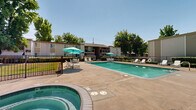 Located at 8079 Sunrise East Way in Citrus Heights, California, this outdoor recreational area is a well-appointed communal amenity space centered around a generously sized swimming pool with clear turquoise water. The pool is complemented by an adjacent circular hot tub/spa, providing residents with a range of aquatic leisure options. The expansive concrete pool deck surrounds the water on all sides, offering ample room for movement and relaxation.

Throughout the deck, multiple white lounge chairs and chaise lounges are arranged for sunbathing, while several seating clusters featuring white tables and chairs are shaded by vibrant teal market umbrellas, providing comfortable spots for outdoor dining or socializing. The entire area is enclosed by a black metal security fence with gated access, ensuring safety and privacy.

The space is framed by mature trees, including tall, slender cypress and broad-canopied shade trees, which provide a natural backdrop and contribute to the overall sense of a lush, sheltered environment. Low-lying landscaping and hedges line portions of the perimeter, adding greenery and visual appeal. The surrounding residential buildings, finished in neutral tan tones, are visible from the pool area. Under the clear California sky, this outdoor space delivers a resort-like atmosphere suited for warm-weather enjoyment.