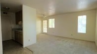 This single-floor space offers a clean, functional layout with an open and airy feel throughout. The main living area is generously sized, finished with soft wall-to-wall carpeting that adds warmth underfoot. Neutral, light-toned walls create a bright and welcoming atmosphere, complemented by natural light streaming in through a large glass-paned door and an adjacent window. These openings provide views of lush greenery outside, connecting the interior to the natural surroundings. A ceiling fan is centrally positioned overhead, offering both practical airflow and a subtle design element. The open layout of the living area allows for flexible furniture arrangement, making it adaptable to a variety of uses. A partial wall divides the living space from an adjoining area, hinting at a defined kitchen or entryway zone just beyond. The overall design is straightforward and understated, prioritizing open space and natural light. The combination of neutral finishes, carpeted floors, and abundant daylight gives the space a calm and comfortable ambiance, well-suited for everyday living.