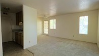 This single-floor space offers a clean, functional layout with an open and airy feel throughout. The main living area is generously sized, finished with soft wall-to-wall carpeting that adds warmth underfoot. Neutral, light-toned walls create a bright and welcoming atmosphere, complemented by natural light streaming in through a large glass-paned door and an adjacent window. These openings provide views of lush greenery outside, connecting the interior to the natural surroundings. A ceiling fan is centrally positioned overhead, offering both practical airflow and a subtle design element. The open layout of the living area allows for flexible furniture arrangement, making it adaptable to a variety of uses. A partial wall divides the living space from an adjoining area, hinting at a defined kitchen or entryway zone just beyond. The overall design is straightforward and understated, prioritizing open space and natural light. The combination of neutral finishes, carpeted floors, and abundant daylight gives the space a calm and comfortable ambiance, well-suited for everyday living.