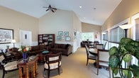 Located at 63 Newfield in Waterbury, this single-story community clubhouse offers a welcoming and thoughtfully appointed gathering space designed for both relaxation and socializing. The interior is defined by its striking vaulted ceilings, which rise dramatically overhead and are complemented by ceiling fans and recessed lighting, creating an airy, open atmosphere throughout.

The main area is divided into distinct functional zones that flow seamlessly into one another. A comfortable lounge area anchors one side of the room, furnished with a large sectional sofa, armchairs, and a coffee table, making it an inviting spot for casual conversation or quiet reading. A well-stocked bookcase lines the wall nearby, adding a warm, library-like touch to the space.

Adjacent to the lounge, a dining area features dark wood tables and upholstered chairs, ideal for communal meals or small gatherings. Large windows along the perimeter flood the space with natural light and offer views of the surrounding greenery, enhancing the sense of openness. Neutral-toned walls and light carpeting throughout contribute to a calm, cohesive aesthetic. Wainscoting detail along the lower walls adds a refined architectural character, lending the space an approachable yet polished ambiance.