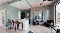 This single-story fitness center is thoughtfully designed to balance active and relaxed spaces. The main lounge area is anchored by floor-to-ceiling windows that frame lush outdoor views of palm trees and landscaped grounds, flooding the interior with natural light. Vaulted ceilings and a ceiling fan enhance the open, airy atmosphere, while light wood-tone flooring runs throughout, providing a clean, cohesive foundation.

A central bar-height counter with a granite surface and sleek bar stools serves as a functional refreshment and gathering area. Adjacent to it, dark cabinetry with a matching granite countertop provides additional storage and workspace, accented by an octagonal wood-framed mirror. Leather seating, including a tufted Chesterfield sofa and armchairs, creates comfortable lounging zones suited for rest and recovery.

The lounge transitions seamlessly into the dedicated workout area, visible through glass-paneled doors. This separation maintains an energetic workout environment while preserving the lounge's calm ambiance. Neutral gray and muted teal wall tones lend a modern, polished character throughout, complemented by curated artwork and thoughtful accent furnishings. A workstation near the windows offers a functional space for administrative or personal use.