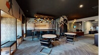 Located at 1200 S Indiana Ave in Chicago's South Loop, this single-floor space delivers a striking, urban-inspired interior that blends lounge comfort with functional coworking amenities. The design is anchored by bold Wall Street-themed murals — featuring iconic street signs, a charging bull silhouette, and large-scale figurative artwork — that lend the space a dynamic, metropolitan character. Dark ceilings contrast with lighter walls, while layered carpet tiles in cool grey tones ground the open layout throughout.

The main lounge area features a mix of seating arrangements, including round white-top tables paired with sleek black-and-gold cantilever chairs, alongside a high-top bar counter with pendant lighting overhead — a cluster of gold-toned drum lights that serve as a dramatic focal point. A tufted cognac leather sofa adds warmth to the lounge corner near a refreshment station outfitted with Marshall-branded equipment, a wall clock, and herringbone tile accents.

A dedicated row of private window nooks provides individual workstations with exterior views, accompanied by artistic travel-themed silhouette murals above. A quieter sitting area features grey wingback chairs, a flat-screen display, hexagonal side tables, and a glass partition etched with a large dial-pad graphic. The overall ambiance is sophisticated yet inviting, ideal for both focused work and casual gatherings.