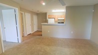 This single-floor space showcases an open-concept design that seamlessly connects the living area to the kitchen. The living area features wall-to-wall beige carpet, contributing to a warm and comfortable atmosphere, while a ceiling fan overhead provides both practical ventilation and a subtle design element.

The kitchen is positioned at a slightly elevated counter level, creating a natural partition between the two spaces without fully closing them off. It is equipped with light wood cabinetry, a white refrigerator, and a gas range, all illuminated by a fluorescent overhead light. The countertop peninsula serves as both a workspace and an informal divider, enhancing the open flow of the layout.

Neutral taupe-toned walls run throughout the space, lending a cohesive and understated aesthetic. Hardwood-style flooring transitions from the kitchen area into a hallway, where two white doors lead to additional rooms, suggesting private spaces such as bedrooms or a bathroom. The overall design emphasizes functionality and simplicity, making it a versatile and move-in ready environment.