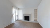 This single-story residence is designed with a clean, neutral palette and a practical layout that flows naturally from room to room. The living area serves as the focal point of the home, featuring a brick-surround wood-burning fireplace painted white, a built-in bookshelf unit ideal for display or storage, and vaulted ceilings that lend a sense of openness. Natural light enters through a sliding glass door with sheer curtains, offering access to an exterior balcony or patio. Soft carpeting extends throughout the living spaces and bedrooms, while wood-look flooring defines the entry, kitchen, and bathroom areas.

The kitchen is thoughtfully appointed with gray cabinetry, white countertops, a double-basin sink, a black dishwasher, and an electric range. A peninsula with a decorative overhead hood and pendant lighting separates the kitchen from the dining area, where a mirrored accent wall reflects light and adds visual depth. The layout opens toward the living room, creating a connected, social atmosphere.

The bedroom is a quiet, carpeted space with a natural light window and a textured ceiling. The bathroom features wood-look flooring, a stone-look vanity countertop, gray cabinetry, and a full tub-shower combination with white tile surround.