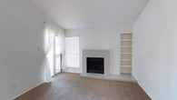 This single-story residence is designed with a clean, neutral palette and a practical layout that flows naturally from room to room. The living area serves as the focal point of the home, featuring a brick-surround wood-burning fireplace painted white, a built-in bookshelf unit ideal for display or storage, and vaulted ceilings that lend a sense of openness. Natural light enters through a sliding glass door with sheer curtains, offering access to an exterior balcony or patio. Soft carpeting extends throughout the living spaces and bedrooms, while wood-look flooring defines the entry, kitchen, and bathroom areas.

The kitchen is thoughtfully appointed with gray cabinetry, white countertops, a double-basin sink, a black dishwasher, and an electric range. A peninsula with a decorative overhead hood and pendant lighting separates the kitchen from the dining area, where a mirrored accent wall reflects light and adds visual depth. The layout opens toward the living room, creating a connected, social atmosphere.

The bedroom is a quiet, carpeted space with a natural light window and a textured ceiling. The bathroom features wood-look flooring, a stone-look vanity countertop, gray cabinetry, and a full tub-shower combination with white tile surround.