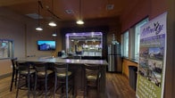 This pub offers a welcoming and well-appointed single-floor layout designed for comfortable dining and socializing. The centerpiece of the space is a wraparound bar illuminated by striking purple LED lighting and pendant lamps, complete with draft taps, glassware displays, and a stainless steel refrigerator. A wall-mounted flat-screen TV is positioned near the bar, adding to the lively atmosphere.

The dining area is generously sized, accommodating a mix of freestanding dark wood tables with cushioned ladder-back chairs and plaid-upholstered booth seating along the walls. Warm golden and terracotta tones on the walls create an inviting ambiance, complemented by light hardwood flooring throughout. Pendant lighting fixtures hang from a tray ceiling with wood trim accents and a tile drop ceiling, providing ample illumination.

Natural light enters through multiple windows fitted with horizontal blinds, offering views of the surrounding area. Decorative vintage-style framed posters, including classic cognac and café advertisements, add character to the walls. Interior pass-through windows connect areas of the space. Track lighting and recessed fixtures provide additional layered lighting, making the pub feel both functional and atmospheric.