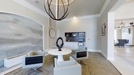 This thoughtfully designed single-floor space exudes a refined, contemporary elegance from the moment you enter. The room is anchored by a striking spherical orb chandelier that serves as a dramatic focal point, casting warm light across the neutral-toned interior. Crown molding and elevated ceilings add a sense of grandeur and architectural polish throughout.

The main gathering area features a sleek round white pedestal table surrounded by modern swivel chairs upholstered in light gray, creating an inviting setting for meetings or casual conversation. A decorative black sculptural centerpiece adds an artistic contrast to the clean, bright furnishings.

Along one wall, a built-in workstation with dark cabinetry and open shelving provides functional storage and workspace, complemented by a compact desktop setup — ideal for administrative or business use. The patterned carpet flooring adds texture and warmth to the otherwise crisp, neutral palette of soft whites and greiges.

A frosted or etched glass partition wall introduces a subtle decorative element, offering both visual interest and a sense of division within the space. Framed artwork on the walls and an octagonal mirror round out the carefully curated décor. Overall, the space balances practicality with sophisticated style, making it well-suited for professional or community use.