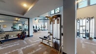 This modern fitness center is designed to support a comprehensive workout experience within a spacious, single-floor layout. The facility features soaring ceilings in key areas, creating an open and airy atmosphere throughout. Large windows line multiple walls, flooding the space with natural light and offering views of the surrounding outdoor greenery.

The cardio zone is anchored by a row of Life Fitness treadmills positioned alongside expansive floor-to-ceiling mirrors that visually enhance the depth of the room. A dedicated strength training area houses a multi-functional cable machine and a functional trainer rig with full cable attachment options, providing versatility for a wide range of resistance exercises. Free weights and adjustable benches are also available, catering to both beginner and advanced users.

Additional cardio equipment, including stationary bikes, is strategically distributed throughout the facility. The patterned carpet flooring with hardwood-tone accents adds a polished, contemporary aesthetic underfoot. Wall-mounted televisions and sanitization stations are thoughtfully integrated throughout the space. The facility benefits from a glass-door entry and multiple access points, ensuring easy flow between exercise zones. The overall design balances functionality with a clean, upscale ambiance suited to daily fitness use.