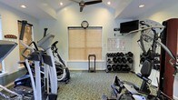 This single-floor fitness room offers a practical and well-equipped space for residents or guests to maintain an active lifestyle. The room features vaulted ceilings with a ceiling fan that promotes air circulation during workouts, contributing to a comfortable exercise environment. Soft green walls and patterned carpeted flooring give the space a welcoming, understated aesthetic. A central window with wooden blinds allows natural light to filter in while providing privacy, and an analog wall clock keeps exercisers on schedule. The cardio area includes at least one elliptical machine and additional equipment positioned along the walls, offering options for low-impact aerobic training. On the opposite side, a multi-tiered dumbbell rack holds a comprehensive range of free weights, catering to various strength training needs. A wall-mounted flat-screen television provides entertainment during workouts, while a bulletin board displays fitness-related charts and informational materials. Recessed lighting supplements the natural light, ensuring the space remains well-lit throughout the day. The overall layout is thoughtfully organized to maximize functionality within the available footprint.