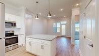 This well-appointed single-floor residence combines modern design with functional living spaces. The heart of the home is an open-concept kitchen and dining area, anchored by a central island topped with speckled granite countertops and crisp white shaker-style cabinetry. Stainless steel appliances, including a refrigerator, range, microwave, and dishwasher, complement the kitchen's clean aesthetic. Pendant lighting with brushed gold fixtures adds warmth and definition to the space. The living and dining areas flow seamlessly from the kitchen, enhanced by crown molding, recessed lighting, and light hardwood-style flooring that runs throughout the main living spaces. Natural light pours in through multiple windows and a glass-paneled door, offering pleasant outdoor views. The bedrooms are carpeted for comfort and feature large windows with blinds, while one room provides convenient access to an in-unit washer and dryer. The bathroom is finished with neutral large-format tile, a white vanity with granite countertop, brushed nickel fixtures, and a well-lit mirror. Additional spaces include a utility closet and versatile flex room with hardwood-style flooring. The overall palette of soft whites and light neutrals gives the entire residence a bright, airy, and inviting atmosphere.