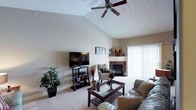 This single-floor space showcases a welcoming living room defined by its striking vaulted ceiling, which creates an impressive sense of height and openness. A ceiling fan is centrally mounted at the peak, providing both functionality and a visual anchor to the soaring roofline. The neutral, warm-toned walls complement the light beige carpeting, contributing to a cohesive and inviting atmosphere throughout.

The living room is thoughtfully arranged around a tile-surround fireplace, which serves as a natural focal point along one wall, complete with a modest mantle ideal for display. A flat-screen television on a dark wood media stand is positioned opposite the seating area, which includes a leather sofa and an upholstered accent chair. A dark wood-and-glass coffee table anchors the seating arrangement, adding an elegant contrast to the soft flooring.

Natural light enters the space through a large window dressed with vertical blinds, brightening the room without overwhelming it. Decorative touches, including potted plants and curated accessories, add warmth and personality. The overall ambiance is comfortable, functional, and well-suited for relaxed living.