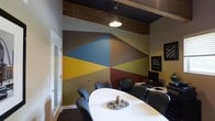 This single-floor space makes a striking first impression with its bold, geometric mural that dominates the main room's focal wall. The mural features large angular shapes in a rich palette of deep blue, mustard yellow, olive green, burgundy, and warm gray, creating a visually dynamic backdrop that sets the tone for the entire space. Overhead, exposed wood beams contrast beautifully against the dark ceiling, lending an industrial yet warm character to the room. A sleek, oval white conference table sits at the center of the room, surrounded by dark leather chairs, making it well-suited for meetings or collaborative work. A pendant light fixture drops from the ceiling, providing focused illumination over the table. Along one wall, a workstation with a monitor and computer setup suggests a functional, professional environment. A frosted glass door adds a touch of modern refinement while maintaining a sense of openness and privacy. The overall ambiance blends contemporary design sensibilities with industrial architectural elements, resulting in a space that feels both creative and purposeful — ideal for a professional office or studio setting.