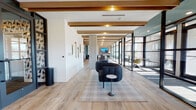 Located at 4015 Craft St in Charlotte, NC, this thoughtfully designed single-floor commercial space blends contemporary aesthetics with functional versatility. Exposed wood beam ceilings add warmth and architectural character throughout, complementing the light-toned hardwood flooring that flows seamlessly across the open layout. The main lounge area features a distinctive curved, channel-tufted sofa anchored on a plaid area rug, with decorative wall-mounted art accents adding a curated, modern touch. A row of floor-to-ceiling black-framed glass walls and doors lines one side of the space, creating a series of private office and meeting rooms that remain visually connected to the broader interior while allowing abundant natural light to pour in. These glass-enclosed offices are furnished with compact tables and ergonomic seating, ideal for focused work or small meetings. A kitchenette or refreshment station features sleek slate-blue cabinetry set against a striking exposed brick accent wall, with a wall-mounted display screen serving as a central media focal point. A long communal work table with bar-height seating provides additional collaboration space. Built-in shelving units and teal accent walls punctuate the interior, adding depth and visual interest. The overall ambiance is polished yet inviting, balancing industrial and contemporary design elements throughout.
