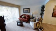 This single-story space showcases a thoughtfully arranged living area designed for comfort and functionality. The room is anchored by plush, light-toned carpeting that extends throughout, contributing to a warm and inviting ambiance. A large sliding glass door fitted with vertical blinds serves as the primary source of natural light, flooding the space with brightness and offering a view of the outdoors.

The living area is furnished with a tufted burgundy loveseat paired with decorative patterned throw pillows, complemented by a neutral-toned armchair draped with a cozy plaid blanket. A round dark-wood coffee table sits at the center, providing a practical yet stylish focal point. A classic table lamp on a slender base adds soft ambient lighting, while a framed landscape painting on the wall introduces a subtle decorative accent. A tall indoor plant in the corner adds a natural, organic element to the space.

The neutral wall tones and white textured ceiling keep the aesthetic clean and understated, allowing the furnishings to stand out. Overall, the space presents a comfortable, livable environment well-suited for relaxed everyday use.