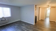 This single-floor space is designed with a straightforward, functional layout that maximizes its footprint. The main living area is finished with dark hardwood-style flooring that runs continuously throughout, providing a cohesive look and easy maintenance. The walls are painted in a soft, neutral grey tone, offering a clean and modern backdrop that works well with a variety of furnishings and décor styles. A textured ceiling adds a subtle architectural detail overhead.

A baseboard heating unit runs along one wall of the living area, providing an efficient heating solution integrated neatly into the space. A partial dividing wall separates the living area from the kitchen zone, where a dark refrigerator is visible, hinting at additional appliances beyond. The kitchen area transitions naturally into a hallway, which leads further into the unit, suggesting additional rooms or storage areas accessible through a visible doorway. The overall layout flows openly from the main living space into the functional areas of the unit, making navigation straightforward. The neutral palette and durable flooring throughout contribute to a clean, move-in-ready ambiance.