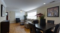 This two-floor home offers a well-designed open-concept main living area that seamlessly connects the living and dining spaces. The living room is anchored by warm hardwood flooring that runs throughout, complemented by crown molding that adds a refined architectural detail along the ceiling perimeter. A ceiling fan with a built-in light fixture provides both comfort and ambient lighting, while large windows fitted with horizontal blinds allow generous natural light to fill the space.

The living area is spacious enough to accommodate a sizable sectional sofa with a chaise lounge, paired with a round coffee table, creating a comfortable and functional seating arrangement. A framed piece of wall art adds a decorative focal point to the neutral-toned walls.

The dining area flows naturally from the living space and features a dark dining table paired with matching chairs, positioned conveniently near a built-in shelving unit with display niches. Recessed lighting in the dining area adds to the layered lighting scheme. The open layout between the living and dining areas creates a cohesive, airy atmosphere ideal for both everyday living and entertaining.