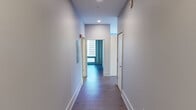 This contemporary single-floor residence showcases an industrial-modern design aesthetic defined by exposed concrete ceilings, warm hardwood flooring, and crisp white walls throughout. The open-concept living and kitchen area serves as the heart of the space, where a spacious island with a light quartz countertop and double sink anchors the kitchen. White cabinetry, stainless steel appliances — including a gas range and full-size refrigerator — and stylish pendant lighting complete the culinary space. A window overlooking the urban skyline fills the kitchen with natural light.

The living area extends toward dramatic floor-to-ceiling windows framed by a structural concrete column, offering sweeping panoramic city views that bring the dynamic urban landscape indoors. The bedroom features dark hardwood floors, an exposed concrete ceiling, dual closets, and a large window framing views of neighboring architecture, complemented by a modern wall sconce.

A long, clean-lined hallway connects the rooms, equipped with recessed lighting and access to additional spaces. The bathroom is a standout feature, boasting a large glass-enclosed walk-in shower finished with gray tile surround, a floating vanity, and light wood-toned tile flooring. The overall ambiance is modern, refined, and distinctly urban.
