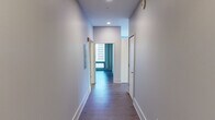 This contemporary single-floor residence showcases an industrial-modern design aesthetic defined by exposed concrete ceilings, warm hardwood flooring, and crisp white walls throughout. The open-concept living and kitchen area serves as the heart of the space, where a spacious island with a light quartz countertop and double sink anchors the kitchen. White cabinetry, stainless steel appliances — including a gas range and full-size refrigerator — and stylish pendant lighting complete the culinary space. A window overlooking the urban skyline fills the kitchen with natural light.

The living area extends toward dramatic floor-to-ceiling windows framed by a structural concrete column, offering sweeping panoramic city views that bring the dynamic urban landscape indoors. The bedroom features dark hardwood floors, an exposed concrete ceiling, dual closets, and a large window framing views of neighboring architecture, complemented by a modern wall sconce.

A long, clean-lined hallway connects the rooms, equipped with recessed lighting and access to additional spaces. The bathroom is a standout feature, boasting a large glass-enclosed walk-in shower finished with gray tile surround, a floating vanity, and light wood-toned tile flooring. The overall ambiance is modern, refined, and distinctly urban.