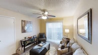 This single-floor living space features a warm and inviting living room designed for everyday comfort and functionality. The room is finished with soft, neutral-toned walls that create a welcoming atmosphere, complemented by wall-to-wall carpeting underfoot. A textured popcorn ceiling adds dimension overhead, while a five-blade ceiling fan with an integrated light fixture provides both air circulation and ambient lighting. A large sliding glass door fitted with vertical blinds serves as a prominent focal point, allowing generous natural light to fill the room while offering access to the outdoors. The space is well-suited for relaxed living, with enough room to accommodate a sofa, accent chairs, and a central ottoman that doubles as a coffee table. A decorative area rug anchors the seating arrangement, adding visual interest to the floor. Along the walls, there is space for a console table with decorative accents, including greenery and small décor items. A floor lamp near the sliding door provides additional task lighting. Overall, the living area offers a practical and comfortable environment with a casual, residential feel.