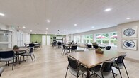 This generously sized single-floor space is designed as a versatile community gathering area, offering ample room for dining, socializing, and group activities. The open layout accommodates numerous tables and chairs arranged throughout, providing flexible seating for a variety of occasions.

The main gathering area features light wood-look flooring, a neutral color palette accented by sage green walls, and a suspended tile ceiling with recessed and flush-mount lighting that ensures consistent, even illumination throughout. Large windows along one wall frame lush outdoor greenery and glimpses of red-umbrella patio seating beyond, filling the interior with abundant natural light and creating a pleasant indoor-outdoor connection.

Framed artwork decorates the walls, contributing a homey, welcoming ambiance. A wall-mounted flat-screen television and a sideboard with decorative accessories add functional and aesthetic detail to one end of the room. Glass-paned interior partitions divide a secondary seating or activity alcove, offering a semi-private space while maintaining visual openness.

A compact kitchenette supports the space, equipped with white cabinetry, a stainless steel refrigerator, a sink with marble-style countertops, and a green tile backsplash. The overall design balances practicality with a warm, inviting atmosphere well-suited for communal use.