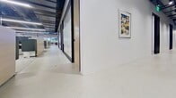 This contemporary office occupies a single floor and offers a well-designed, functional workspace that balances collaborative and focused work environments. The layout includes an open-plan workstation area outfitted with height-adjustable desks, ergonomic seating, and panel dividers that provide individual focus while maintaining an open feel. Exposed structural ceilings with steel beams and visible ductwork give the space an industrial character, complemented by sleek linear LED lighting throughout.

A dedicated conference room features a central meeting table surrounded by ergonomic chairs, a wall-mounted multi-panel display, a whiteboard, and neutral carpeting underfoot — creating a polished setting for presentations and collaboration. The corridor connecting the spaces is lined with full-height glass partitions framed in black, lending a clean, modern aesthetic and allowing natural light to flow freely through the interior.

Large perimeter windows in the open workspace area frame lush, tree-filled outdoor views, flooding the floor with natural light and creating a bright, energizing atmosphere. White walls display framed artwork, adding a touch of personality to the professional environment. The overall design strikes a balance between industrial and refined, making it a versatile and inviting workplace.